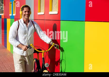 Uomo sorridente in camicia bianca che posa con una bicicletta gialla contro una parete colorata con motivi geometrici Foto Stock