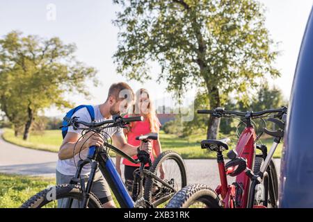Il giovane uomo e una donna si preparano per la bicicletta fuoristrada, abbattendo mountain bike elettriche dal portabiciclette sul camper. Foto Stock