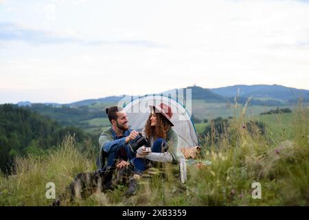 Coppia di viaggiatori che trascorrono del tempo all'aperto, dormendo in un riparo per tende. Giovane turista seduto nella natura, bevendo caffè, ammirando il tramonto. Vacanze estive Foto Stock