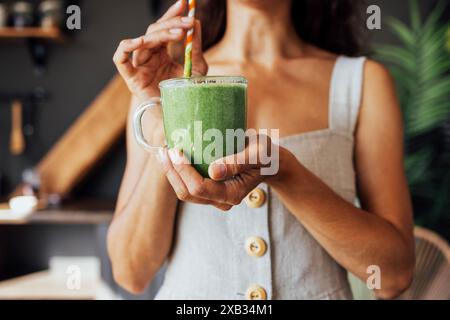 Una tazza trasparente con un frullato verde nelle mani di una giovane donna. Una ragazza in un prendisole di lino beve una bevanda fresca fatta di erbe e verdure Foto Stock