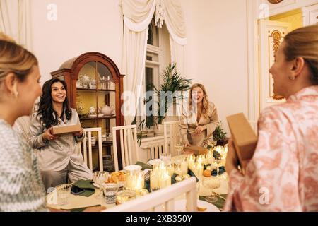 Giovani donne attraenti in pigiama ridono e scambiano regali al tavolo delle feste. Ragazze affascinanti si divertono insieme a una festa di Capodanno. Il ricco st Foto Stock