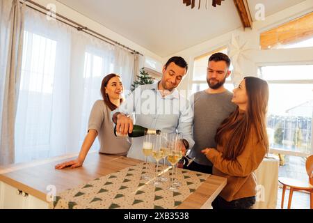 Un giovane sorridente versa champagne nei bicchieri nella sala da pranzo della casa. Due attraenti coppie sposate festeggiano la vigilia di Natale. Amici felici ce Foto Stock