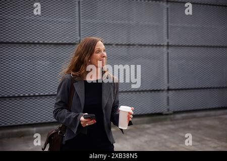 Donna d'affari sorridente con tazza usa e getta e smartphone sul sentiero Foto Stock