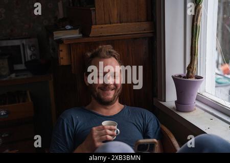 Uomo felice che beve un caffè mentre usa lo smartphone vicino alla finestra a casa Foto Stock