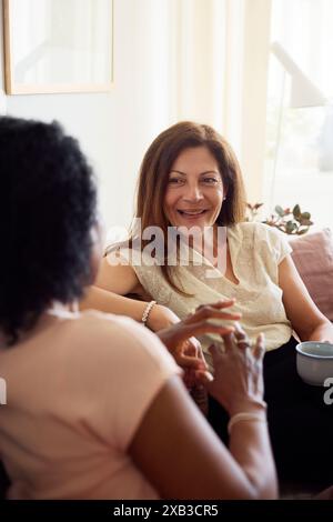 Donna sorridente con una tazza di caffè che parla con un'amica mentre trascorri il tempo libero a casa Foto Stock