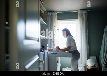 Vista laterale della giovane madre che cambia il pannolino del bambino stando accanto alla finestra Foto Stock