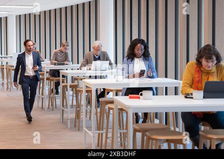 Uomo d'affari che cammina accanto ai colleghi seduti a un tavolo nella caffetteria dell'ufficio Foto Stock