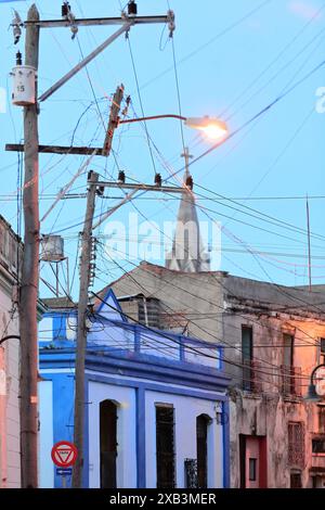 366 groviglio di cavi elettrici collegati a pali di pubblica utilità con il campanile della chiesa di Sagrado Corazon sullo sfondo, Calle padre Olallo Street. Camaguey-Cuba. Foto Stock