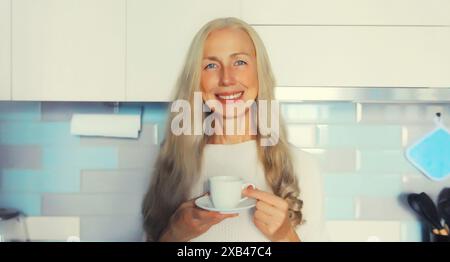 Ritratto di una donna dai capelli grigi sorridente di mezza età che si gusta una gustosa tazza di caffè o tè caldo in cucina la mattina presto a casa Foto Stock