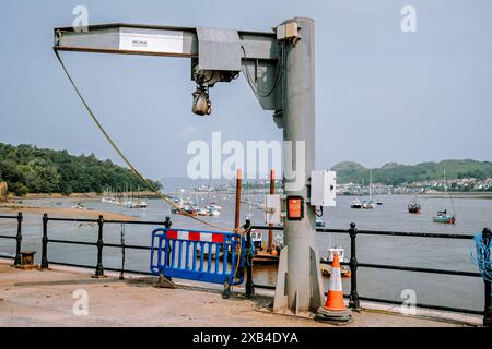 Conwy Wales Regno Unito 06-01-2024. Gru portuale e barriere di sicurezza sul lungomare con barche ancorate in lontananza sotto il cielo limpido Foto Stock