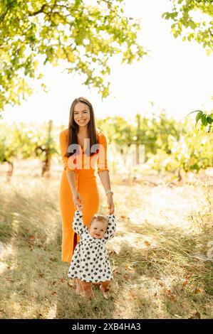 Una donna con un abito arancione e un bambino con pois di polka godendosi una giornata di sole in un vigneto, pieno di amore e felicità Foto Stock