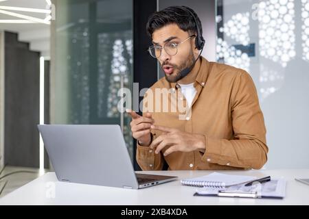 Un giovane indiano serio siede in ufficio alla scrivania con una cuffia e guarda scioccato lo schermo del laptop, legge le notizie, parla durante una videochiamata. Foto Stock