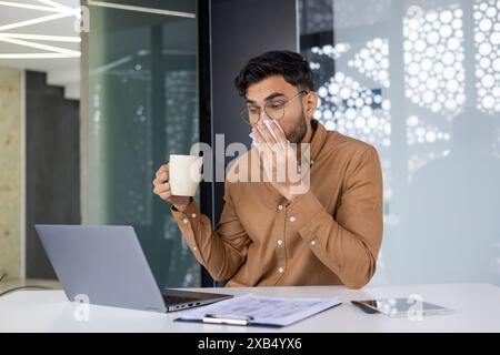 Un giovane indiano malato seduto in ufficio alla scrivania con un portatile, si pulisce il naso che cola con un tovagliolo e tiene in mano una tazza di bevanda calda e medicine. Foto Stock