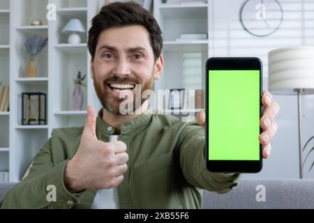 Ritratto ravvicinato di un giovane a casa, seduto sul divano e che mostra alla fotocamera un modello verde di un telefono cellulare e un segnale di un dito. Foto Stock