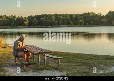 Un anziano anziano seduto a un tavolo da picnic che guarda il suo cellulare mentre il sole tramonta. Foto Stock