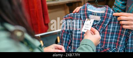 Striscione di un'etichetta irriconoscibile per il cliente femminile prezzo della camicia a quadri nel negozio di seconda mano Foto Stock