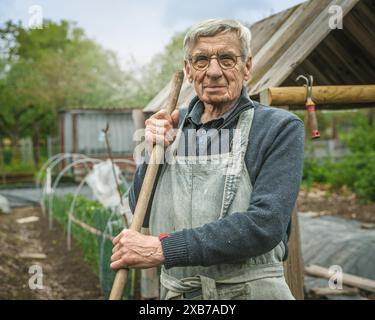 Uomo anziano che indossa un maglione e un grembiule, appoggiato su una pala mentre lavora in un orto, che rappresenta il giardinaggio e le attività all'aperto. Foto Stock