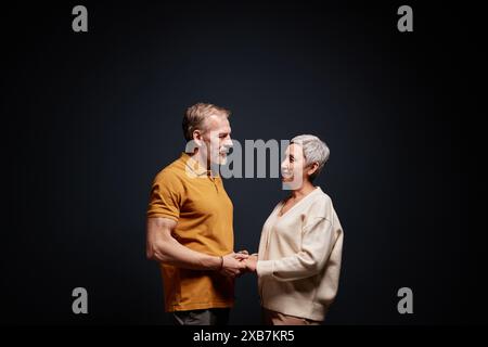 Immagine laterale minima di una coppia senior che tiene per mano e si guarda l'un l'altro su sfondo nero in studio, spazio di copia Foto Stock