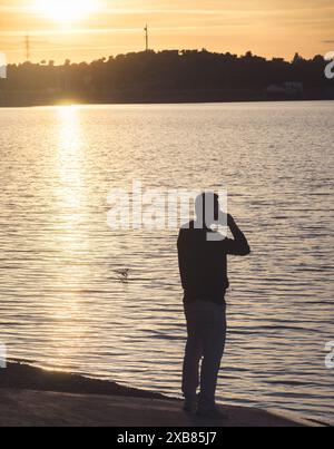 Un uomo è in piedi in un lago, a parlare al cellulare. Il sole tramonta sullo sfondo, proiettando un caldo bagliore sull'acqua. Foto Stock