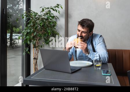 Giovane uomo d'affari affamato durante la pausa pranzo seduto in caffetteria a mangiare hamburger e a usare il computer portatile per lavorare online. Gli amanti degli hamburger, gli uomini d'affari mangiano e. Foto Stock