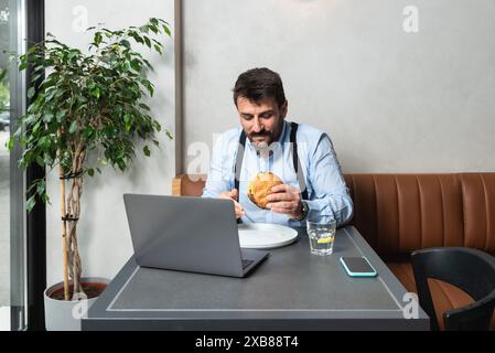 Giovane uomo d'affari affamato durante la pausa pranzo seduto in caffetteria a mangiare hamburger e a usare il computer portatile per lavorare online. Gli amanti degli hamburger, gli uomini d'affari mangiano e. Foto Stock