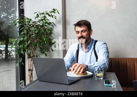 Giovane uomo d'affari affamato durante la pausa pranzo seduto in caffetteria a mangiare hamburger e a usare il computer portatile per lavorare online. Gli amanti degli hamburger, gli uomini d'affari mangiano e. Foto Stock