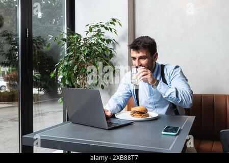 Giovane uomo d'affari affamato durante la pausa pranzo seduto in caffetteria a mangiare hamburger e a usare il computer portatile per lavorare online. Gli amanti degli hamburger, gli uomini d'affari mangiano e. Foto Stock