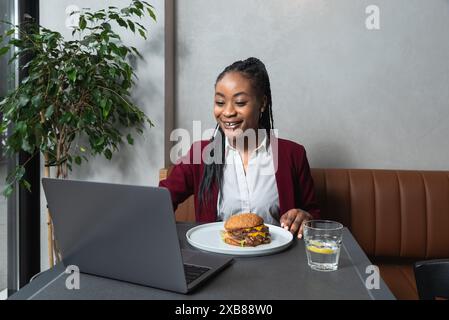 Giovane donna d'affari afroamericana affamata durante la pausa pranzo seduta in caffetteria a mangiare hamburger e a usare il computer portatile per lavorare online. Il cameriere amante del hamburger Foto Stock