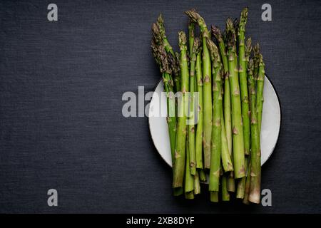 Lancette di asparagi fresche disposte su un piatto bianco su uno sfondo grigio scuro in tessuto. Foto Stock