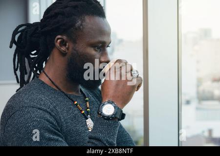 L'uomo africano che guarda attraverso la finestra dell'ufficio, bevendo un caffè e pensando indossa un orologio, una collana e anelli, ha una barba, dreadlock e.. Foto Stock