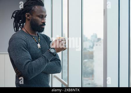 Adulto lavoratore d'ufficio africano uomo è in piedi vicino alla finestra pensando alle strategie aziendali. L'uomo indossa un maglione, un orologio e una collana è lo Foto Stock