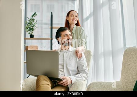 Una donna rossa e un uomo barbuto siedono vicino a una sedia, ingannati nell'uso di un portatile insieme nel loro moderno appartamento. Foto Stock