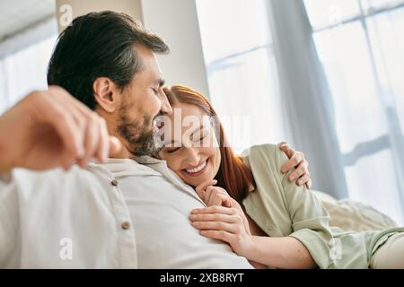 Una donna rossa e un uomo barbuto condividono un caldo abbraccio seduto su un comodo divano nel loro moderno appartamento. Foto Stock