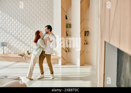 Una bella coppia adulta, una donna rossa e un uomo barbuto che ballano gioiosamente nel loro moderno salotto. Foto Stock