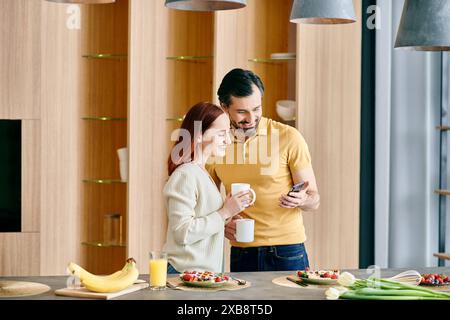 Una donna rossa e un uomo barbuto guardano il loro telefono, in piedi in una cucina moderna, godendo del tempo di qualità a casa. Foto Stock