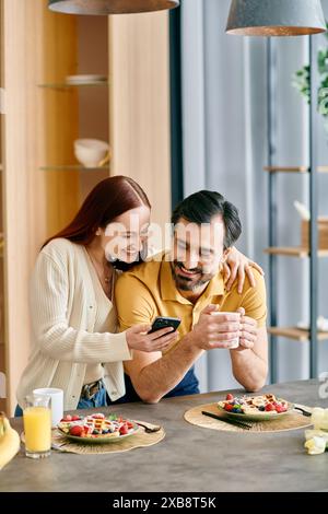 Una donna rossa e un uomo barbuto siedono a un tavolo da cucina, invischiati nei loro telefoni, mentre si godono la colazione insieme. Foto Stock
