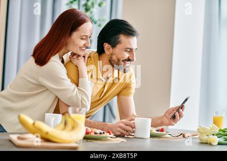 Una bella coppia adulta, una donna rossa e un uomo barbuto, si siedono a un tavolo invischiato nei loro telefoni, godendosi del tempo di qualità insieme a casa. Foto Stock