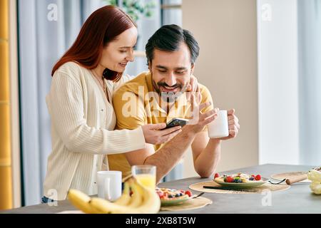 Una donna rossa e un uomo barbuto sono concentrati sui loro telefoni mentre si gusta la colazione insieme in un appartamento moderno. Foto Stock
