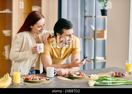 Una donna rossa e un uomo barbuto gusteranno la colazione mentre sono circondati dai loro telefoni in un moderno appartamento. Foto Stock