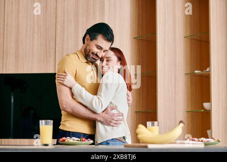 Una donna rossa e un uomo barbuto condividono un caldo abbraccio in una cucina moderna, ridendo e adorando il momento insieme. Foto Stock