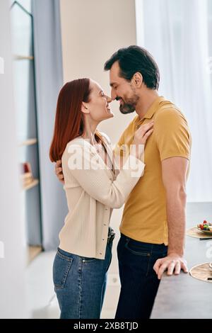 Una donna rossa e un uomo barbuto che condividono un bacio appassionato in una cucina moderna, esprimendo amore e connessione. Foto Stock
