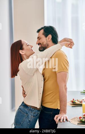Una coppia adulta, una donna rossa e un uomo barbuto condividono un tenero abbraccio nella loro cucina moderna, esprimendo amore e vicinanza. Foto Stock