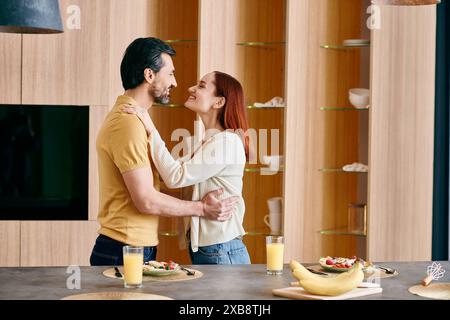 Una donna rossa e un uomo barbuto condividono un tenero abbraccio in una cucina moderna, circondata da calore e affetto. Foto Stock
