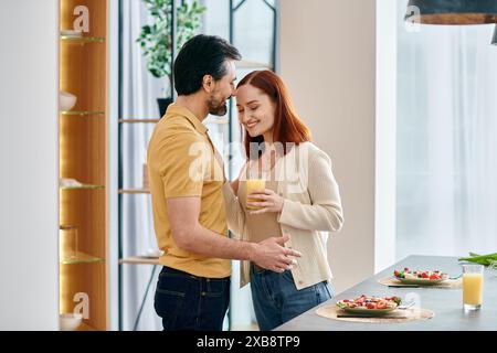 Una donna rossa e un uomo barbuto si trovano in una cucina moderna, ognuno con un bicchiere di succo d'arancia. Foto Stock