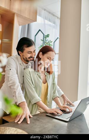 Una donna rossa e un uomo barbuto si sono immersi nello schermo di un notebook mentre si trovano nella loro cucina moderna. Foto Stock