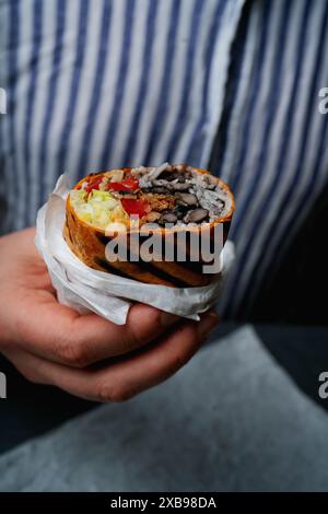 Immagini ravvicinate mostrano una persona che tiene un burrito arrotolato e grigliato, rivelando il ripieno di riso, fagioli, pomodori, lattuga e carne, contro una strada Foto Stock