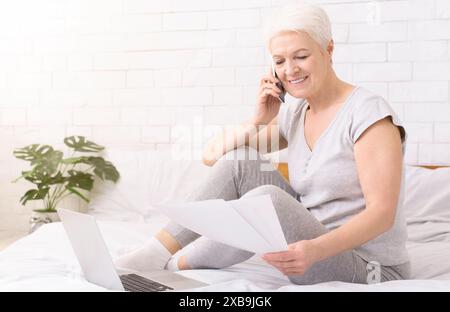 Donna matura sorridente che lavora da casa sul letto durante il giorno Foto Stock