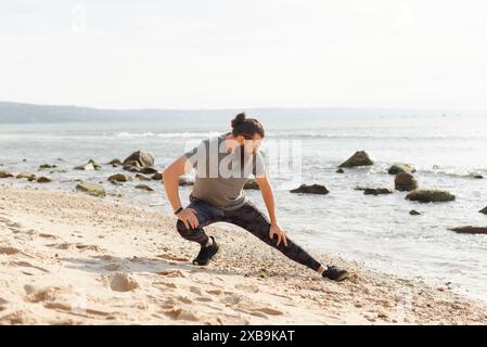 L'uomo che si allunga su una spiaggia sabbiosa con una costa rocciosa sullo sfondo, si concentra sul fitness e sul relax. Foto Stock