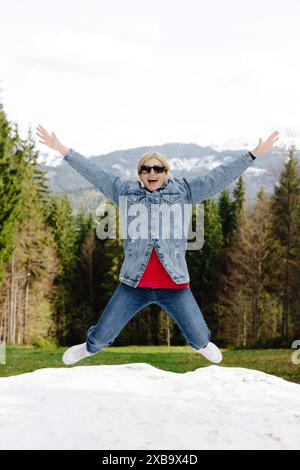 Una donna che indossa una giacca blu in denim e un jeans salta in aria, braccia allungate, di fronte a un paesaggio innevato di montagna. Ha un'espressione felice Foto Stock