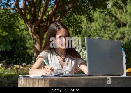 Bella ragazza del college che prende appunti mentre interagisce con il suo portatile nel campus. Giovane studente concentrato, tecnologia, inizio delle lezioni, vita accademica Foto Stock